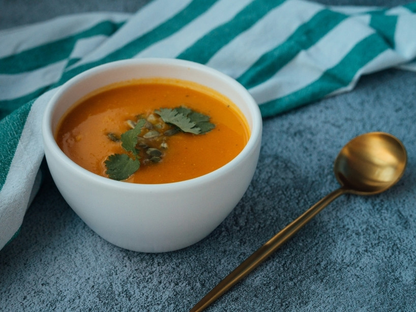 A bowl of soup on a table next to a spoon and a linen napkin.