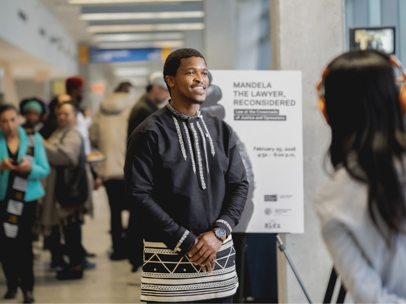 Siyabulela Mandela pictured wearing a traditional black South African tunic with white embroidery. He is being interviewed on camera as event guests mingle in the foyer at the Sears Atrium, Toronto Metropolitan University.