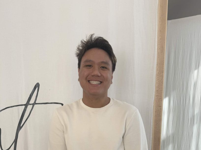 Third-year Marketing Management student Alec Leano smiles at the camera, wearing a cream-colored long-sleeved shirt and black pants, with hands in pockets. They stand in front of a white backdrop with black lettering and a wooden frame to the right.