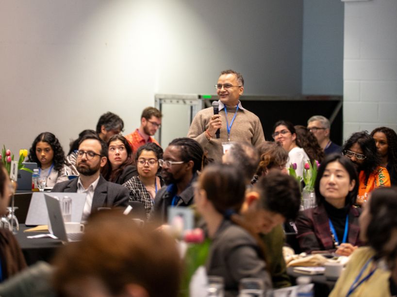A man holding a microphone speaks to a diverse audience seated at tables in a conference room.