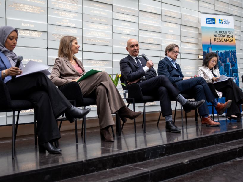 Five panelists sit on chairs on a stage in front of a wall with inscribed text at the launch of the global migration institute. A banner behind them reads "The Home for Migration Research."