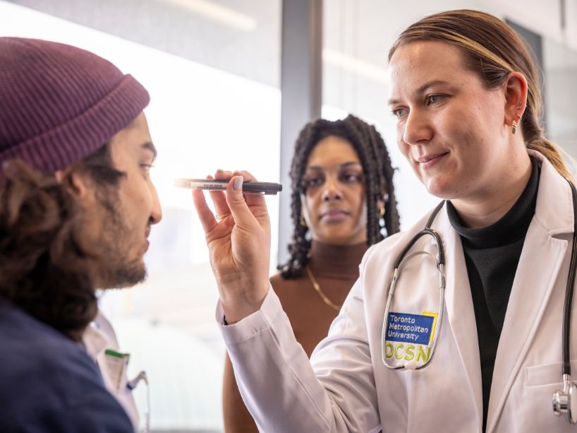 A young woman holds a medical instrument up toward the face of a male patient, as another woman looks on.