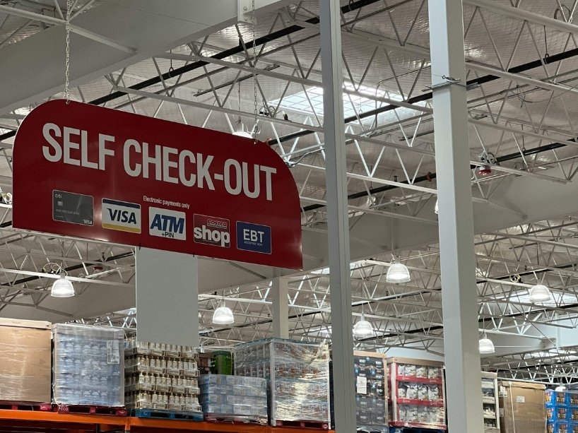 A grocery store with a sign hanging from the rafters indicating the location of the self-checkout.