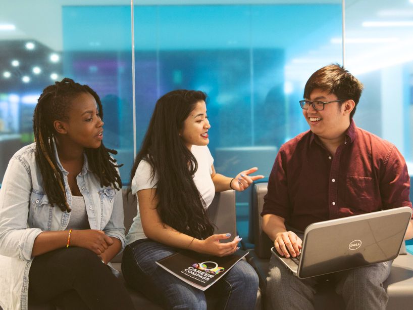 Three students in discussion while seated in front of a blue window.