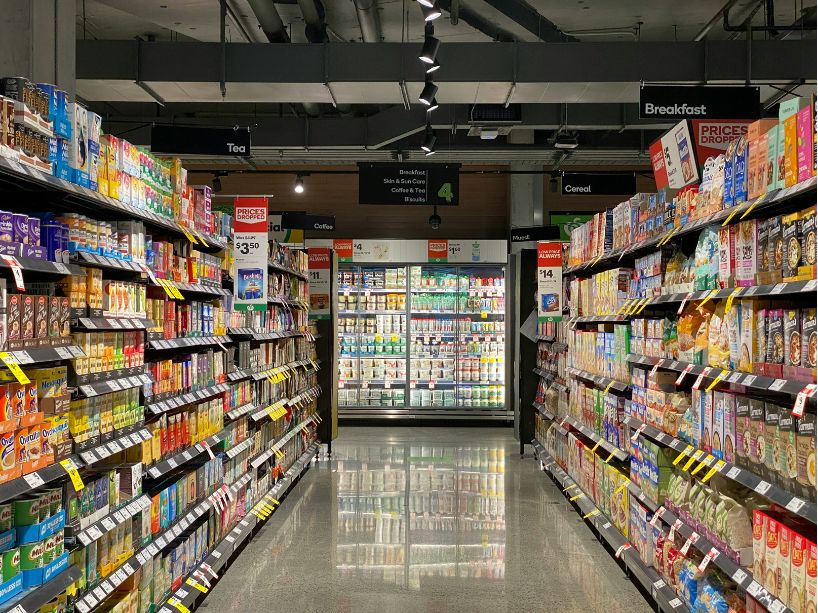 A wide aisle in a supermarket with shelves stocked with breakfast cereals, tea, coffee, and other groceries. Refrigerated units are visible at the end of the aisle.