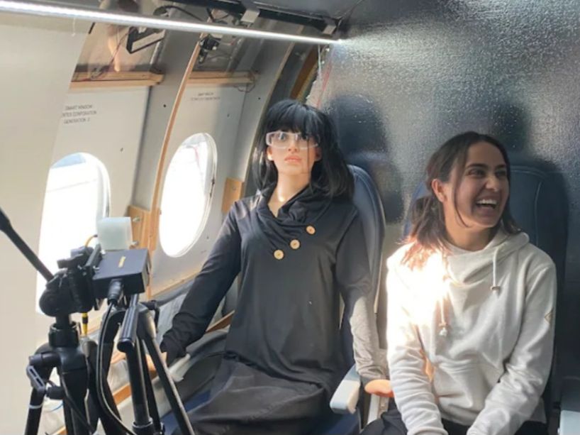 Kritika Vashishtha seated in a mock airplane cabin, next to a research mannequin wearing glasses with sensors measuring light.