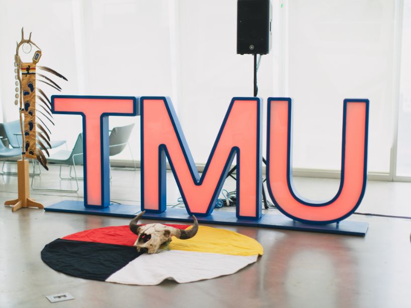 An Indigenous staff with feathers standing vertically beside a large red TMU light-up sign. On the floor, there is an Indigenous medicine wheel with a buffalo/bison skull on top.