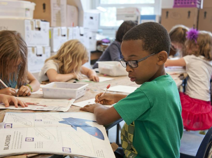 A young boy sits at a table in a classroom writing something on paper and looking at a book, while other kids work around him.