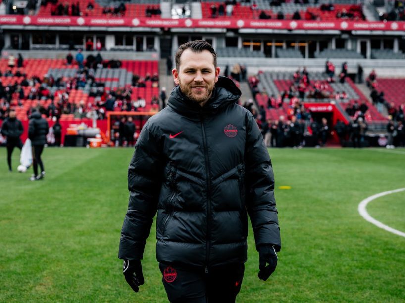 A young man in a black soccer jacket and pants walks across the field looking at the camera.