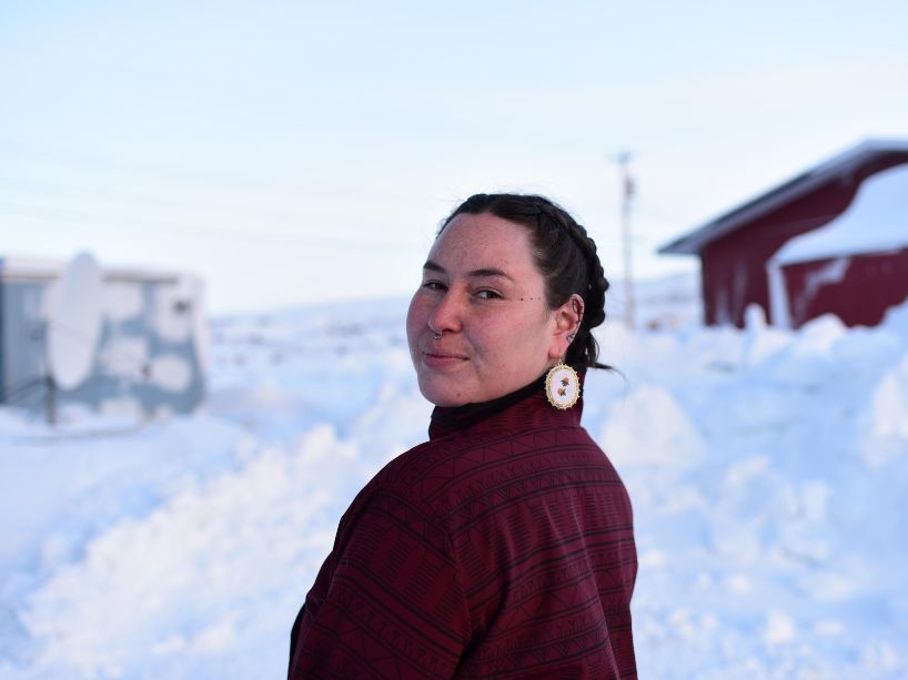 Paige Kimiksana Kreps looks back at the camera framed by two outbuildings in landscape of deep snow.