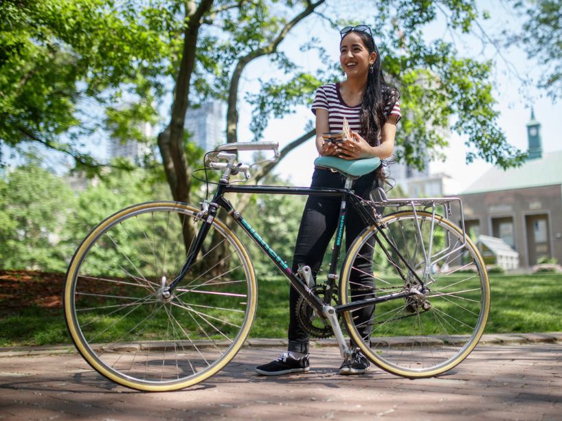 A TMU student stands behind a black and teal bike in the quad.