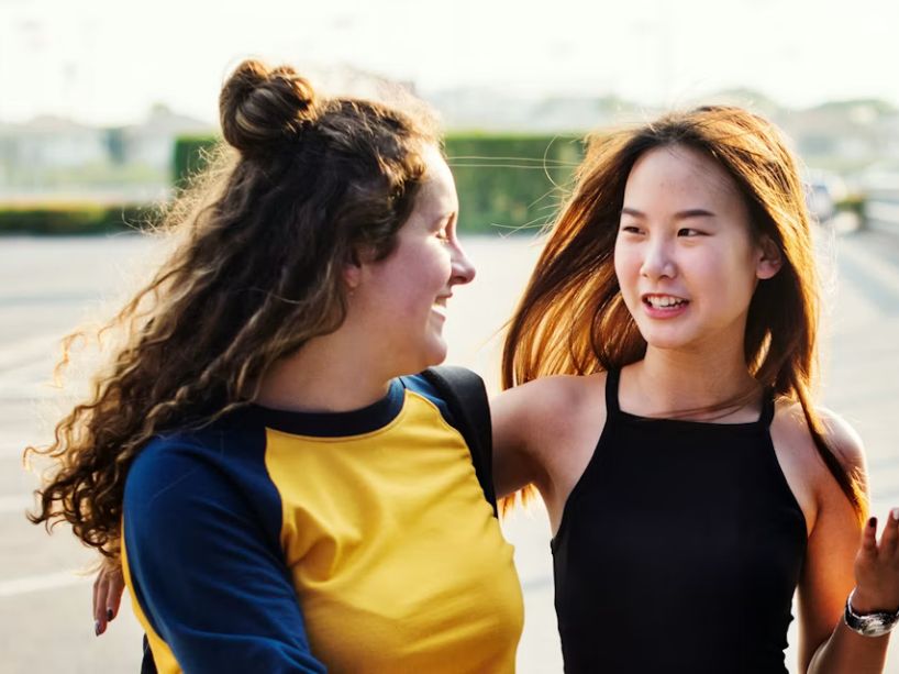 Two friends walking outdoors along a city walkway, smiling and chatting in warm sunlight with modern buildings and railings in the background.
