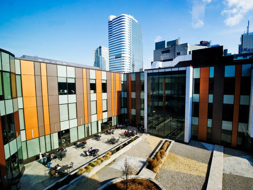 Courtyard between modern office buildings with patterned glass and brick panels, rows of chairs along the walls, a gravel and concrete walkway, and tall city skyscrapers under a blue sky.