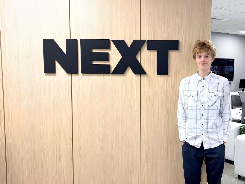 Jake Briand stands to the right of a sign for the NEXT accelerator