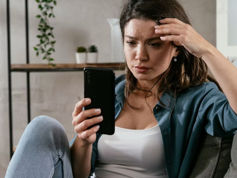 A person seated on a gray couch at home, holding a smartphone in one hand and covering their forehead with the other, looking stressed or unwell in a bright living room with plants.