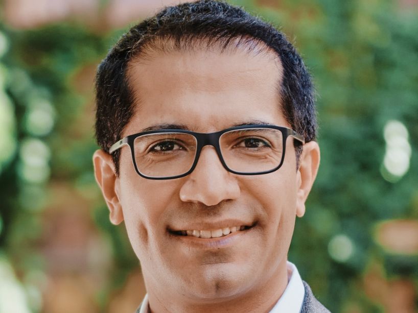 Smiling person wearing black rectangular glasses and a gray blazer over a white shirt, photographed outdoors with a blurred green background.
