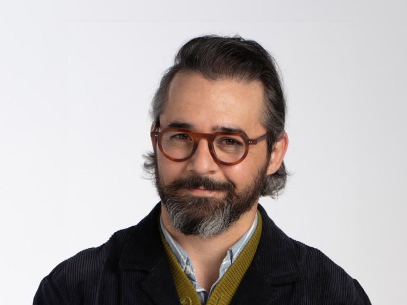 Portrait of a person with dark hair and a full beard, wearing round brown glasses, a dark sweater, and a mustard-yellow collared layer against a plain light background.