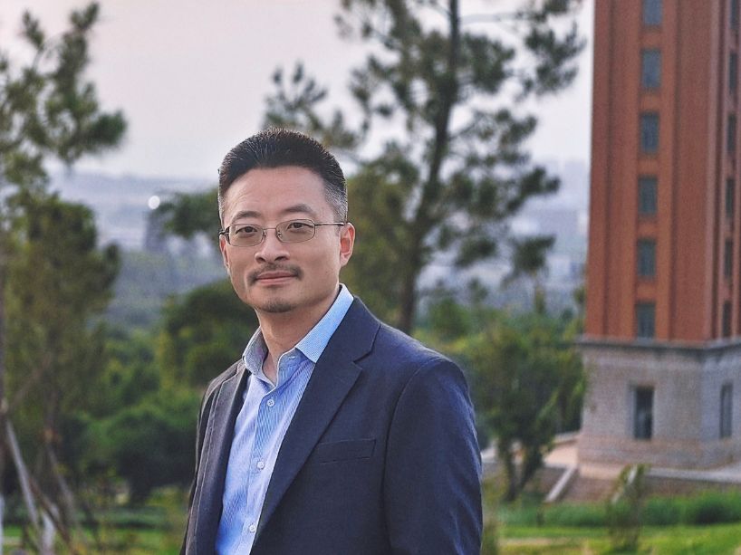 A man in a blue dress shirt and dark blazer stands outdoors in a garden with purple flowers and tall trees, with a building and distant hills in the background.