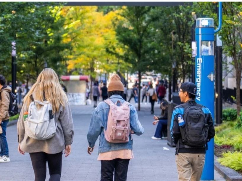 People walk along a tree-lined campus path with backpacks, passing benches on the left and a blue signpost near the center under bright autumn light.