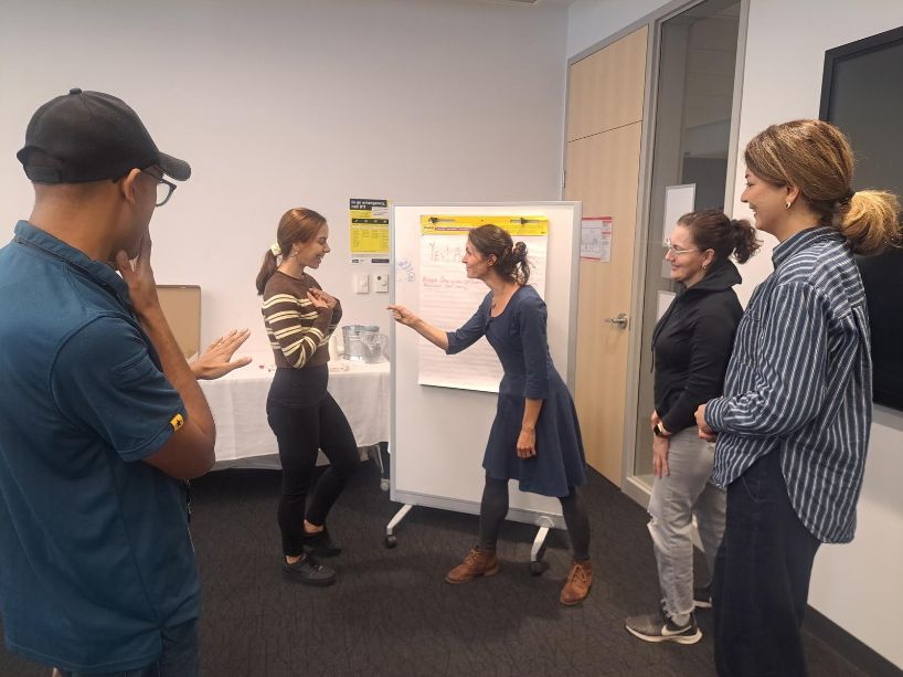 Group of people standing in a training room, watching a presenter point at a whiteboard and discussing printed charts on an easel.