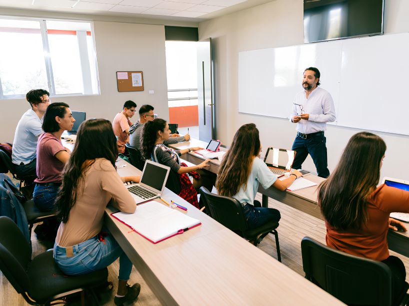 A trainer stands at the front of a bright classroom, speaking to a group of people seated at desks with laptops, while one person writes or gestures during a workshop.