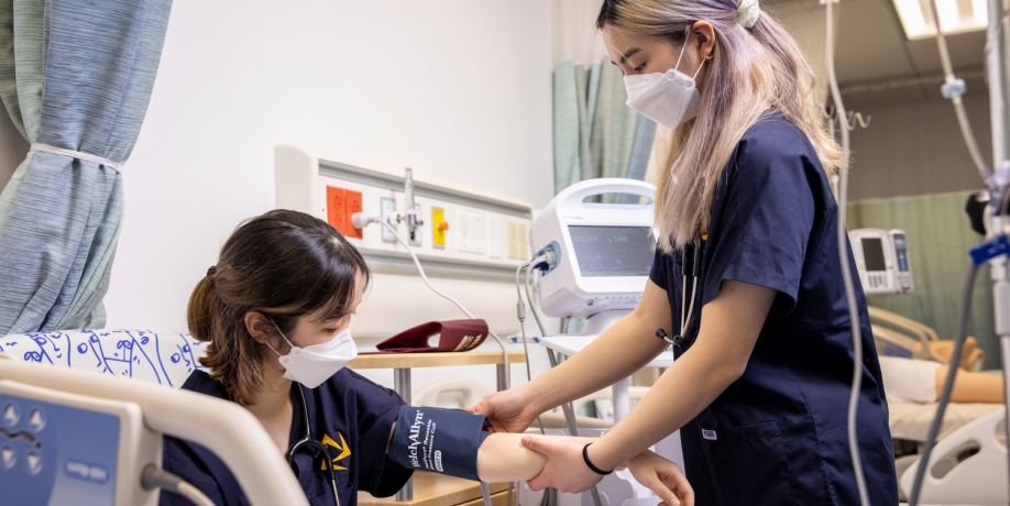 Two masked students in the lab practising measuring blood pressure with a cuff