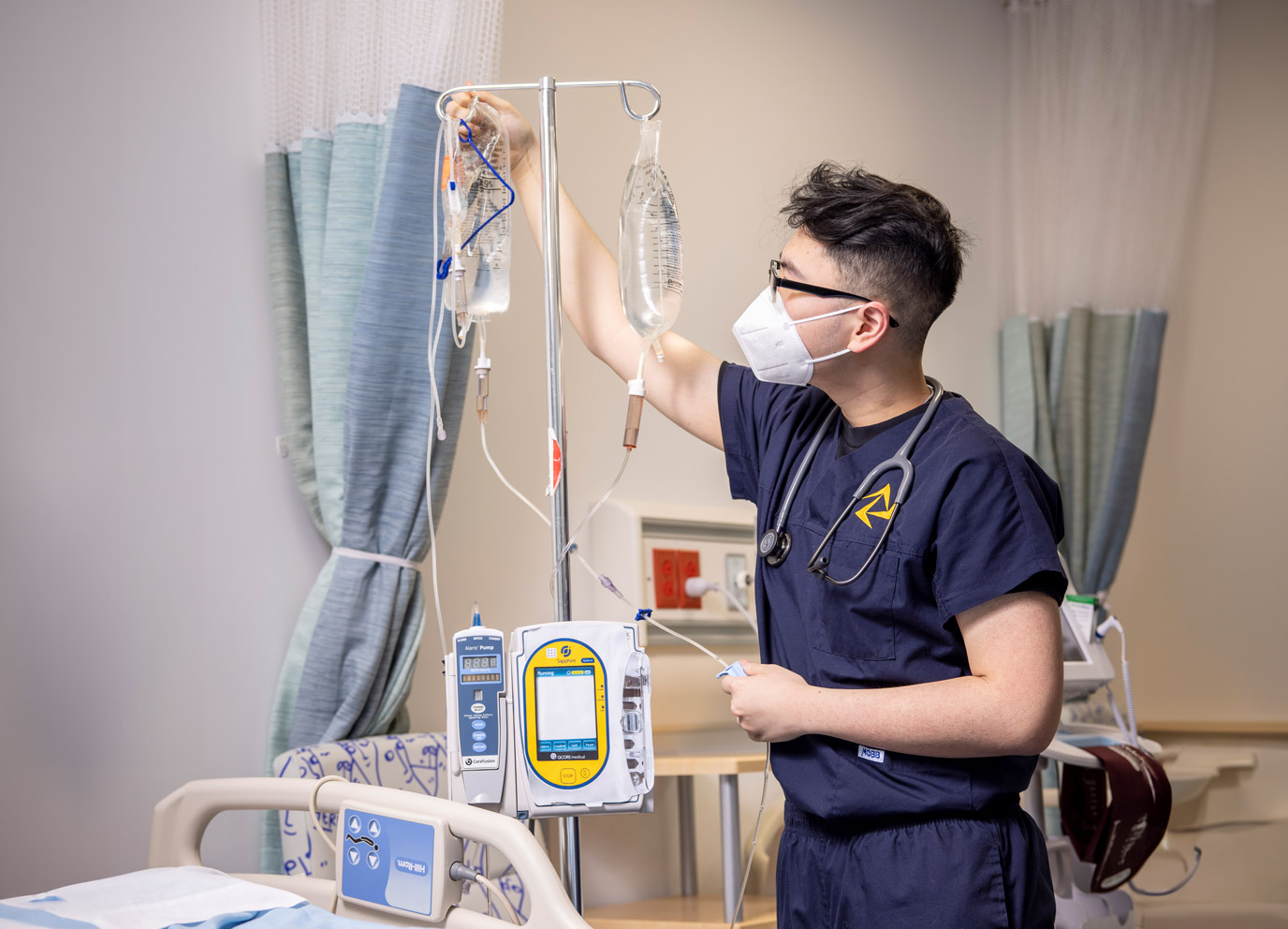 A Collaborative Nursing Program student in scrubs and a face mask checks the fluid levels in an IV bag in a medical training lab.