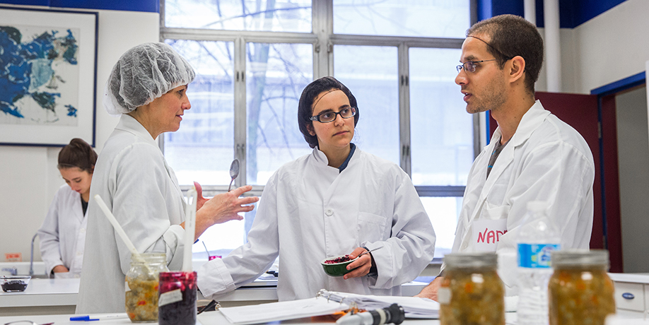 Three Nutrition students in lab coats and hair nets talking amongst themselves in lab