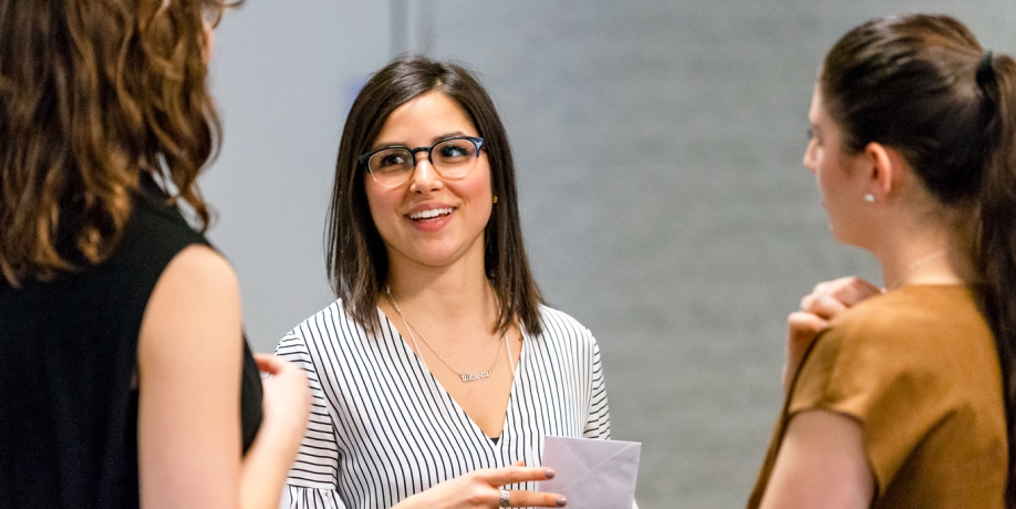 Standing students converse in a group centered on a woman holding a white envelope 