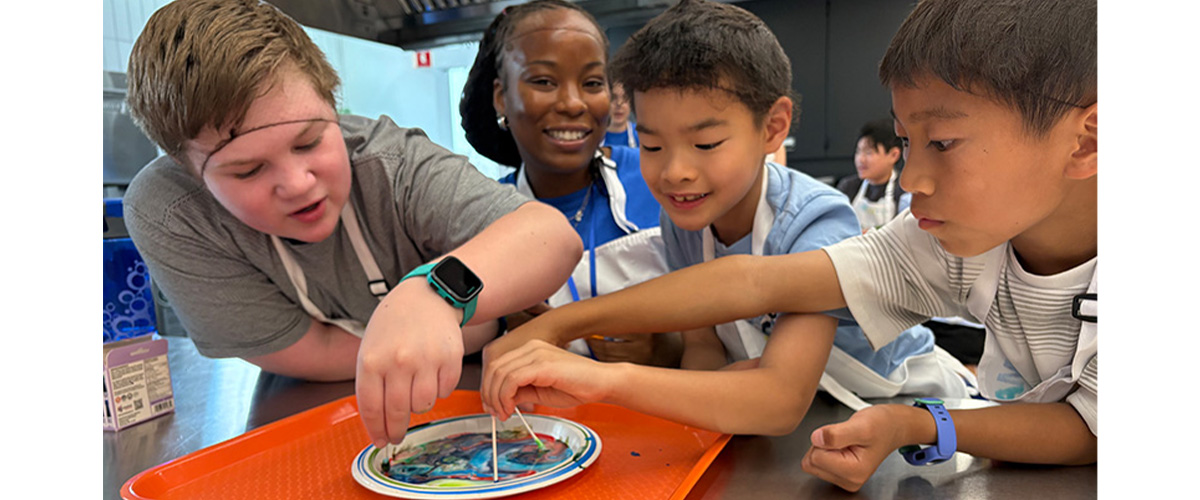 Three children mix food colouring on a plate with an instructor smiling at the camera
