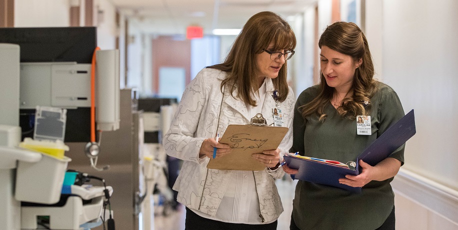 Two women in hospital hallway reviewing notes on clipboard and in binder