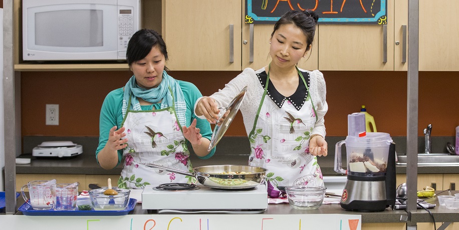 Nutrition students preparing East Asian food in kitchen for TMU food and cultural festival. A sign reading 'East Asia' hangs behind them.