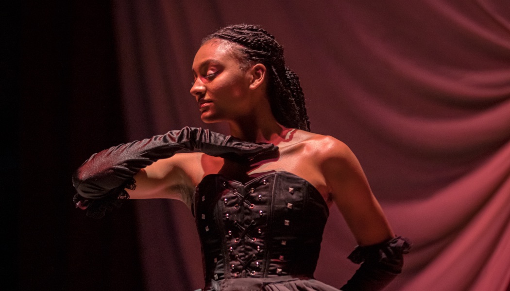 A student performer in a black corset and gloves poses gracefully on stage.