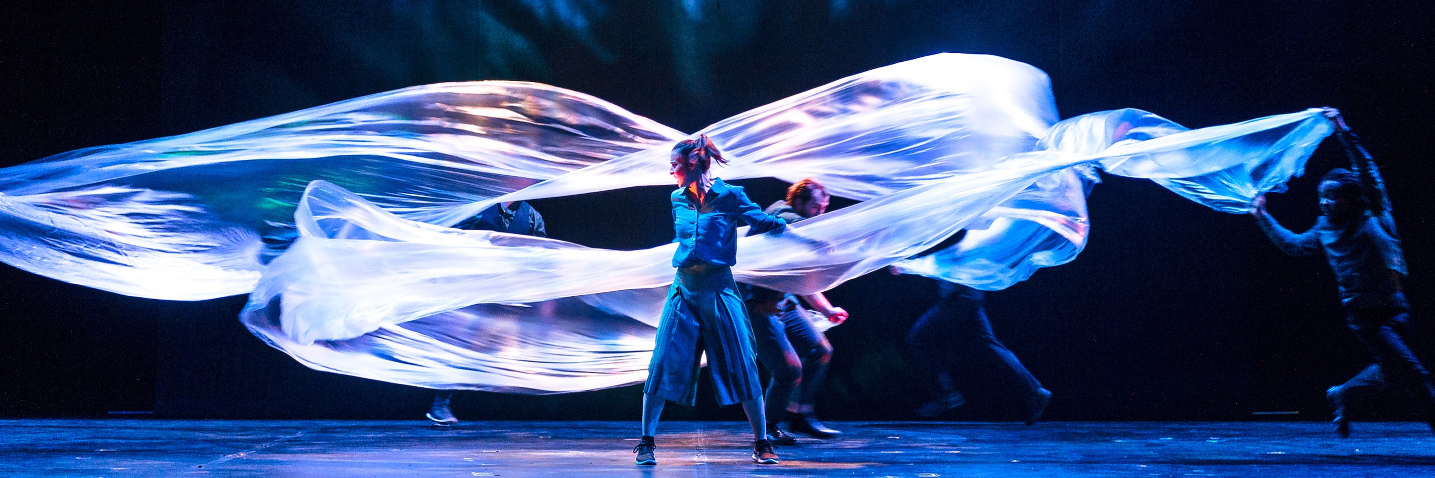 Dancers move across a stage with large, flowing white fabrics, creating an ethereal effect.