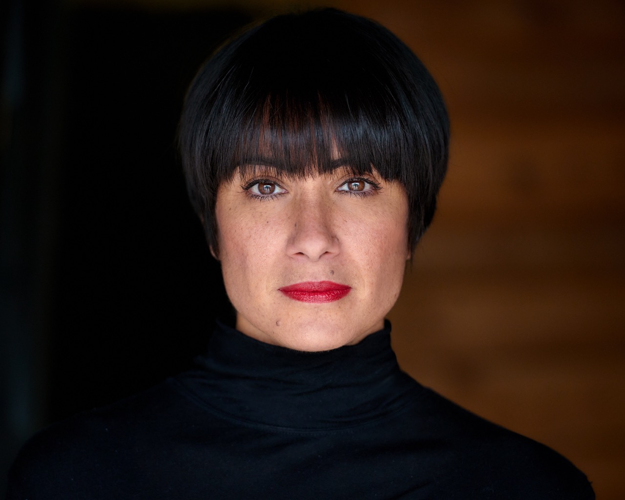 headshot of Anisa Tejpar. she is against a maroon back ground, has short black hair and bangs, she looks at the camera with pursed lips