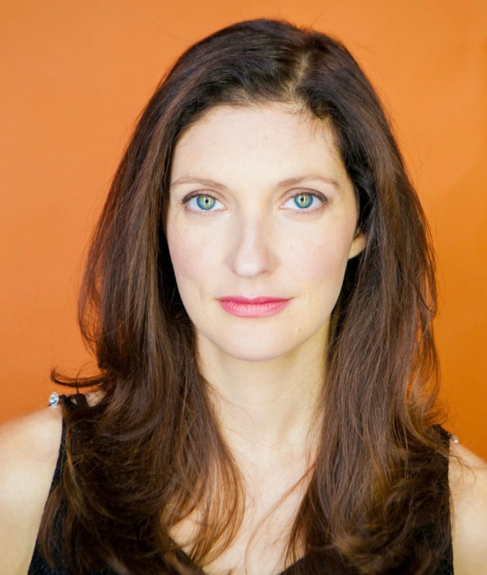 Laura stares intently at the camera with a bright orange backdrop behind her. 