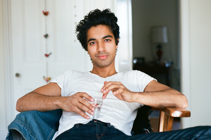Photo of Ishan Dave, he is in a white tshirt and a laid back pose smirking at the camera