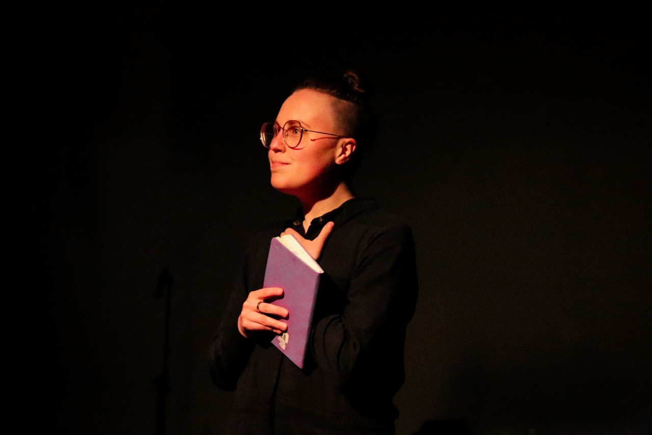photo of sadie-epstien fine. they are in a dark room with a single spotlight looking into the distance holding a purple book