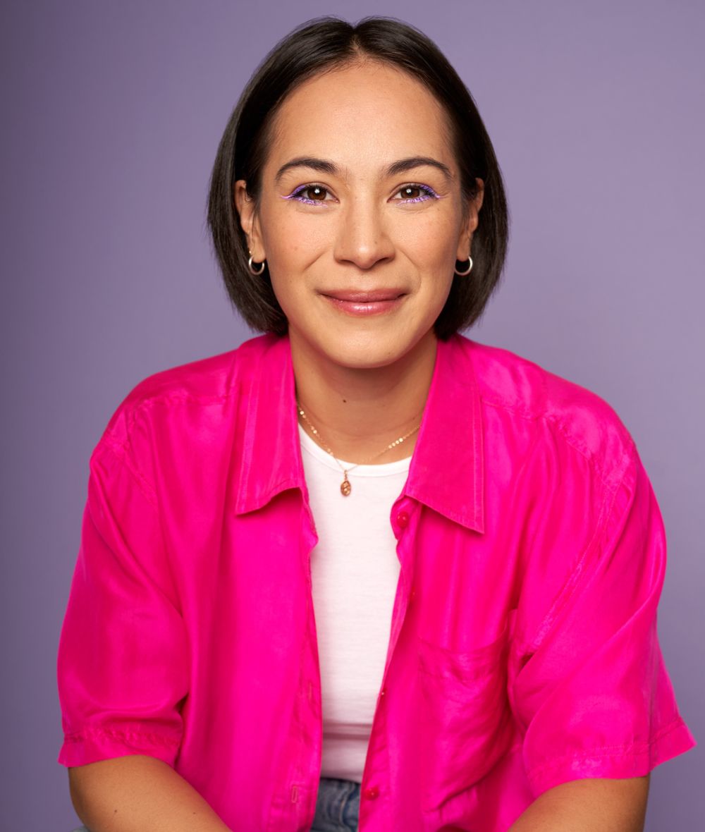 Femme person of Filipinx-German descent in her early 30s sitting casually in front of a lavender background, their hands clasped, leaning forward with kind eyes warmly smiling into camera. They're wearing a silky hot pink short-sleeve button up open over a white tanktop and light blue jeans with simple jewelry. Photo credit: Pascal Lamothe-Kipnes