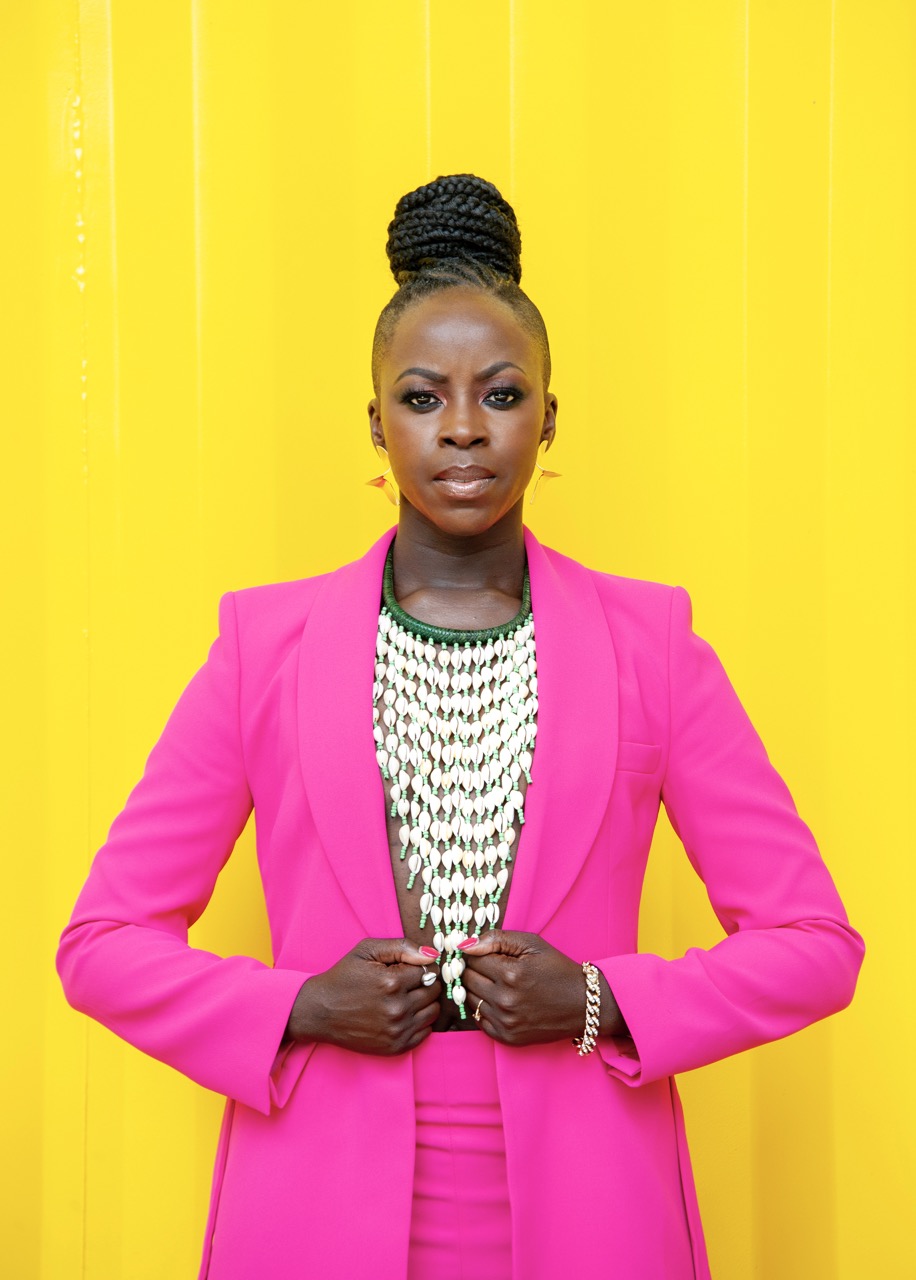 Esie stands strongly against a bright yellow background staring intensely forward. She is wearing a bright pink suit. 