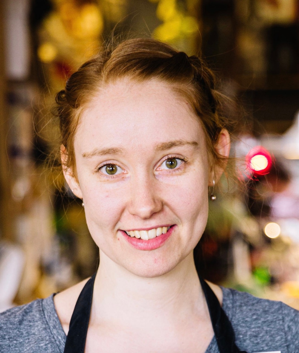 headshot of bronwyn pel, she smiles at the camera, her hair is up, and she is against a multicolour background