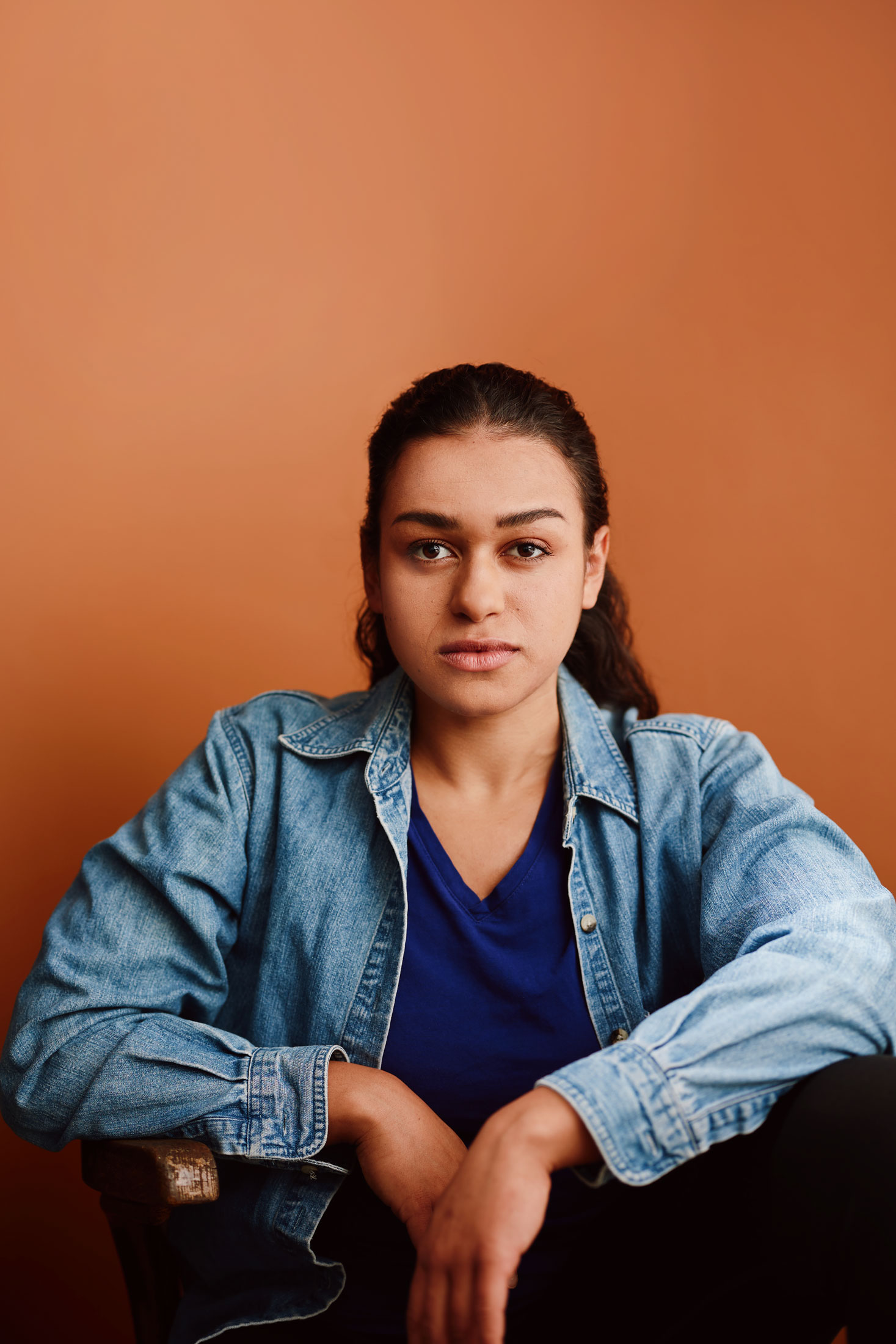 Autumn stares down the camra. She is seated on the ground, resting elbows on her knees, with her arms falling in. She wears a jean jacket, adn her dark hair is in a low ponytail.