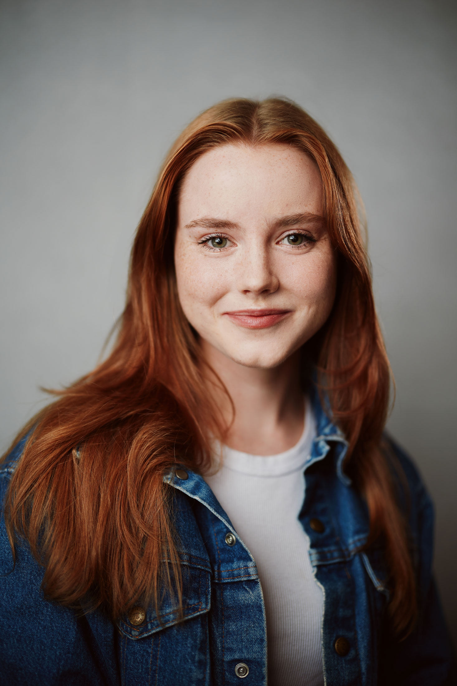 Olivia smiles with a closed mouth. She wears a jean jacket and her red hair is parted in the middle and falls in front of her shoulders.