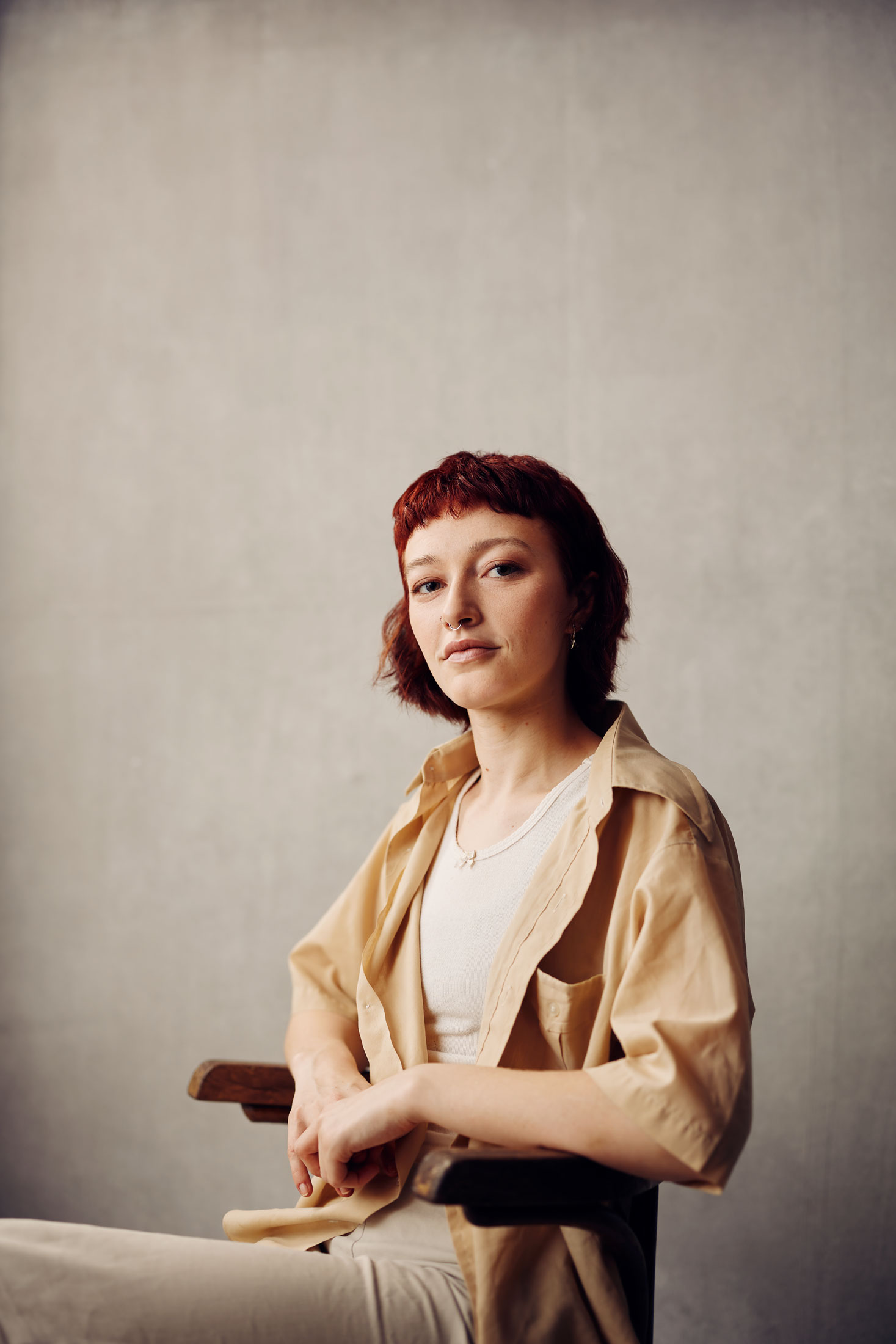 Holly leans back resting their arms on the arms of a chair. They have a short red bob, with a trim fringe. Their pale yellow button up reveals a white top.