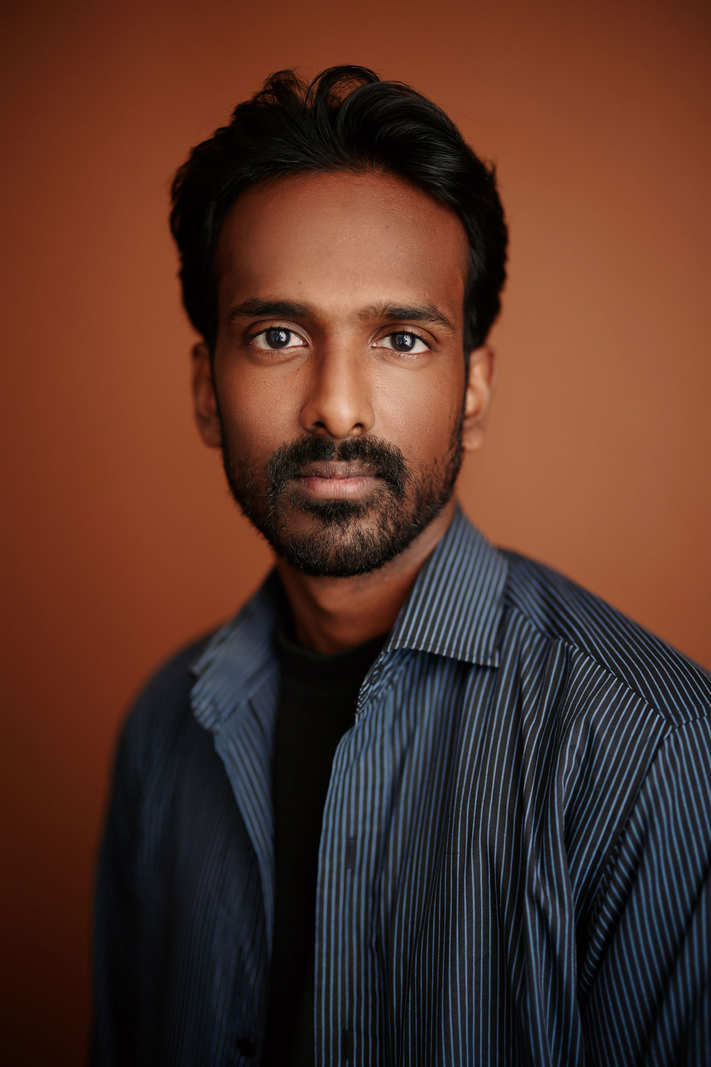 Ben looks at the camera pensively. His blue button up reveals a black shirt underneath. His dark beard is trimmed close to his face.