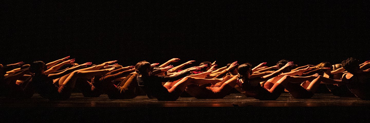 Dancers on a dark stage crouch in unison lit by an ominous orange glow