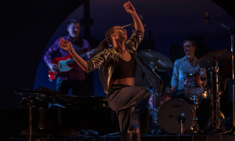 Dancer tosses her head backwards in movement with a jazz band behind her. 