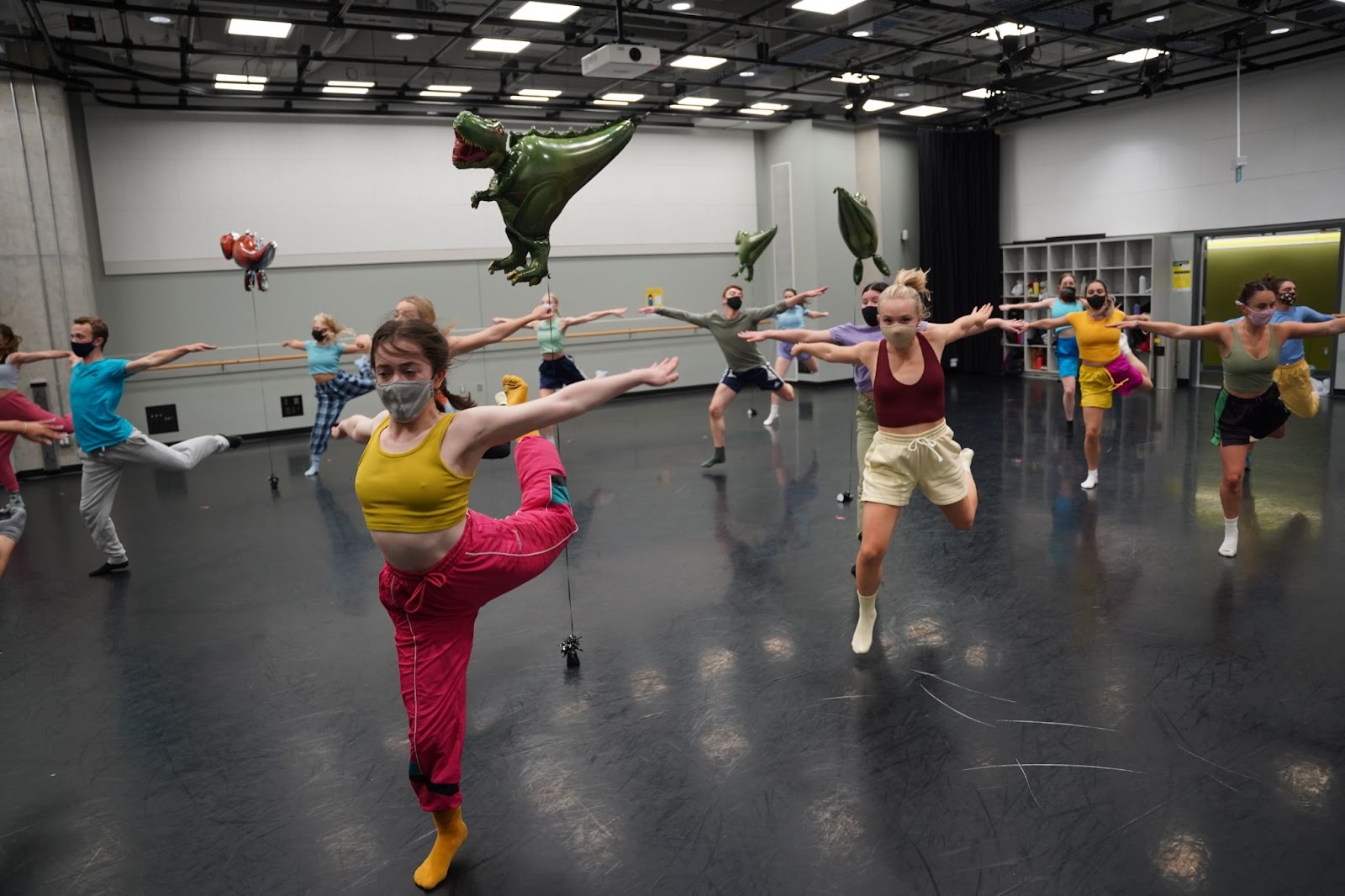 In a studio with dinosaur helium balloons, dancers jump forward onto one leg with their arms thrown behind them