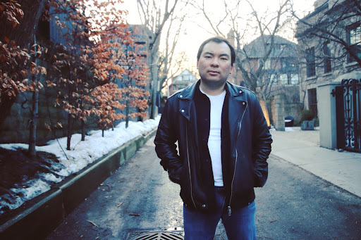 Aaron stands on a street in winter with his hands in the pockets of his leather jacket 