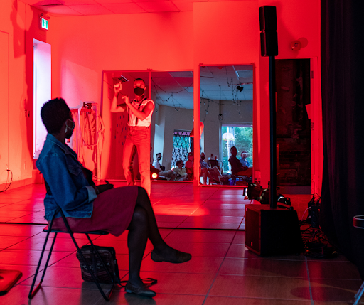 A person sits with crossed legs in a room with red light as another looks on